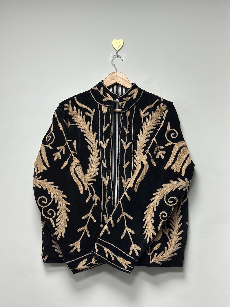 May include: A black jacket featuring intricate gold-coloured embroidery in a leaf and vine pattern. The garment has a high collar, long sleeves, and a front opening that reveals a black and white striped lining. It is displayed on a wooden hanger.
