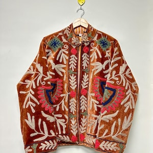 Puede incluir: Una chaqueta de terciopelo color naranja quemado con intrincados bordados en tonos crema, rojo, azul y verde. La prenda tiene cuello alto, mangas largas y una abertura frontal, decorada con patrones florales y de hojas en toda la superficie.