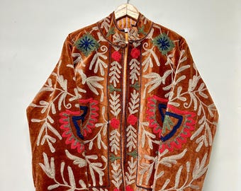 May include: A burnt orange velvet jacket featuring intricate embroidery in shades of cream, red, blue, and green. The garment has a high collar, long sleeves, and a front opening, decorated with floral and leaf patterns across the entire surface.