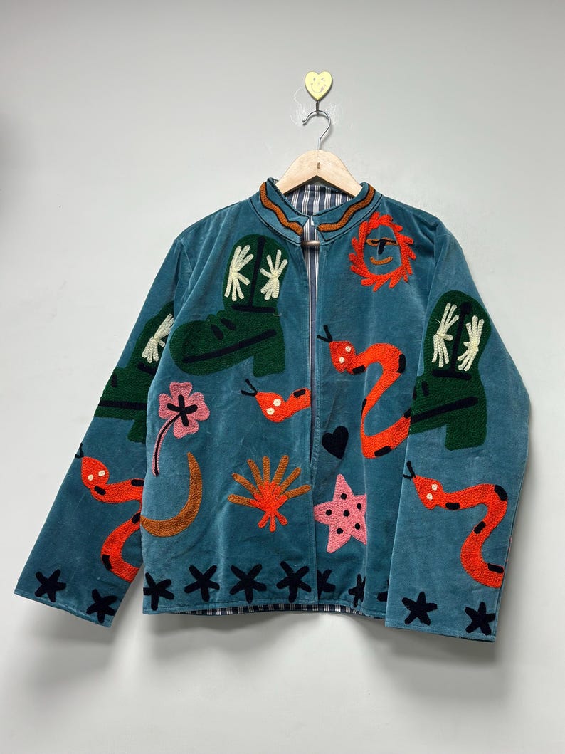 May include: Teal velvet jacket with embroidered designs. Features include green cowboy boots, orange snakes, pink flowers, and black star accents. The jacket has a zip closure and a striped collar.