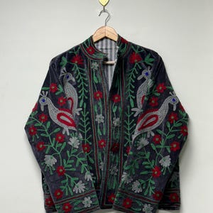 May include: A dark gray velvet jacket featuring intricate embroidery of stylized birds and floral patterns in vibrant red, green, and silver thread. The jacket has a mandarin collar, long sleeves, and a front opening, hanging on a wooden hanger.