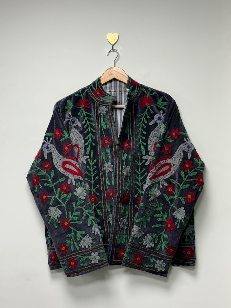 May include: A dark gray velvet jacket featuring intricate embroidery of stylized birds and floral patterns in vibrant red, green, and silver thread. The jacket has a mandarin collar, long sleeves, and a front opening, hanging on a wooden hanger.
