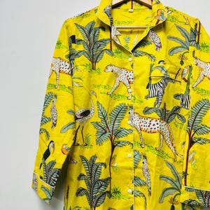 Puede incluir: Camisa de pijama amarilla con botones y estampado de tem&aacute;tica selv&aacute;tica. El dise&ntilde;o presenta leopardos, cebras, p&aacute;jaros y palmeras. La camisa de manga larga tiene ribetes blancos y est&aacute; colgada de una percha de madera.