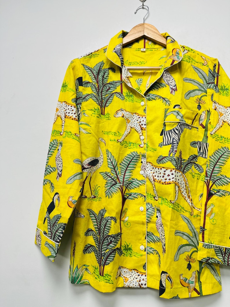 Puede incluir: Camisa de pijama amarilla con botones y estampado de tem&aacute;tica selv&aacute;tica. El dise&ntilde;o presenta leopardos, cebras, p&aacute;jaros y palmeras. La camisa de manga larga tiene ribetes blancos y est&aacute; colgada de una percha de madera.