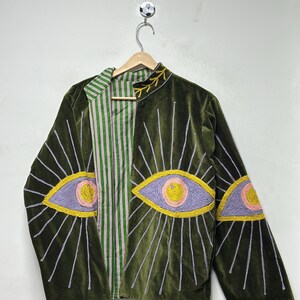 K&ouml;nnte beinhalten: Eine olivgr&uuml;ne Samtjacke mit gestickten Augenmotiven auf der Vorderseite und den &Auml;rmeln. Die Augen sind in lavendelfarbenem, rosafarbenem und gelbem Garn mit strahlenf&ouml;rmigen Linien dargestellt. Das Innenfutter zeigt ein gr&uuml;n-wei&szlig;-rotes Streifenmuster.
