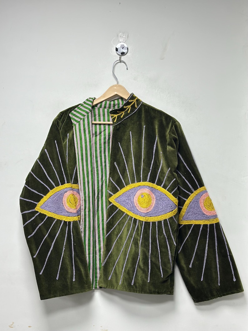 K&ouml;nnte beinhalten: Eine olivgr&uuml;ne Samtjacke mit gestickten Augenmotiven auf der Vorderseite und den &Auml;rmeln. Die Augen sind in lavendelfarbenem, rosafarbenem und gelbem Garn mit strahlenf&ouml;rmigen Linien dargestellt. Das Innenfutter zeigt ein gr&uuml;n-wei&szlig;-rotes Streifenmuster.