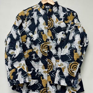 May include: A quilted jacket with a black background and a repeating pattern of white and gold cranes and circular designs. The jacket has a mandarin collar and button closure. The sleeves are long and the jacket is hanging on a wooden hanger.