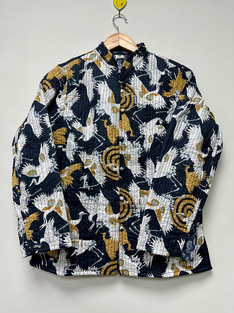 May include: A quilted jacket with a black background and a repeating pattern of white and gold cranes and circular designs. The jacket has a mandarin collar and button closure. The sleeves are long and the jacket is hanging on a wooden hanger.