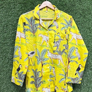 Puede incluir: Camisa amarilla con botones y un estampado de jungla vibrante. El dise&ntilde;o presenta leopardos, cebras, tucanes y palmeras. La camisa tiene mangas largas y ribete blanco en los pu&ntilde;os y el cuello.