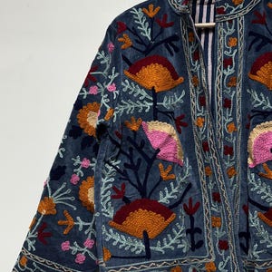 May include: A blue jacket featuring intricate floral embroidery in shades of orange, pink, and dark red. The garment has a high collar, long sleeves, and a front opening that reveals a blue and white striped lining on the interior fabric.