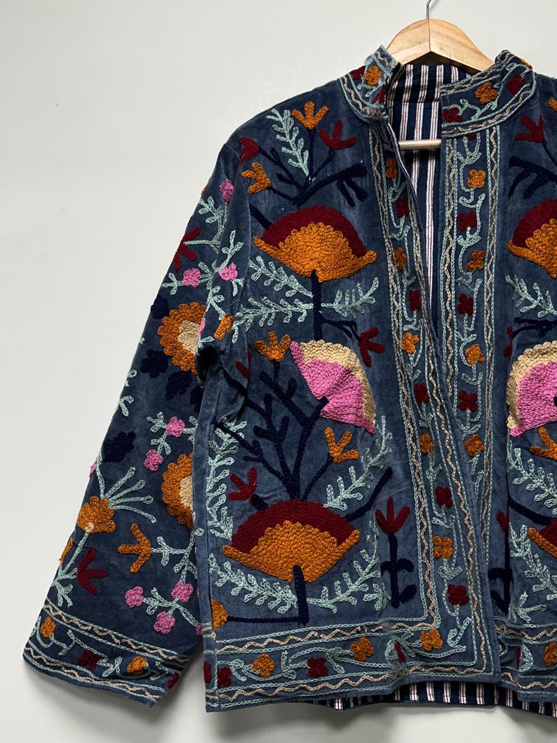 May include: A blue jacket featuring intricate floral embroidery in shades of orange, pink, and dark red. The garment has a high collar, long sleeves, and a front opening that reveals a blue and white striped lining on the interior fabric.