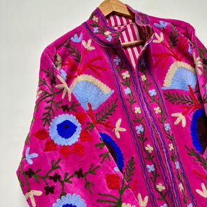 K&ouml;nnte beinhalten: Eine leuchtend pinke Samtjacke mit aufwendigen, mehrfarbigen Blumenstickereien in Blau-, Rot-, Gr&uuml;n- und Cremet&ouml;nen. Die Jacke hat einen Stehkragen, lange &Auml;rmel und ein sichtbares Innenfutter mit einem wei&szlig;-pinken vertikalen Streifenmuster.