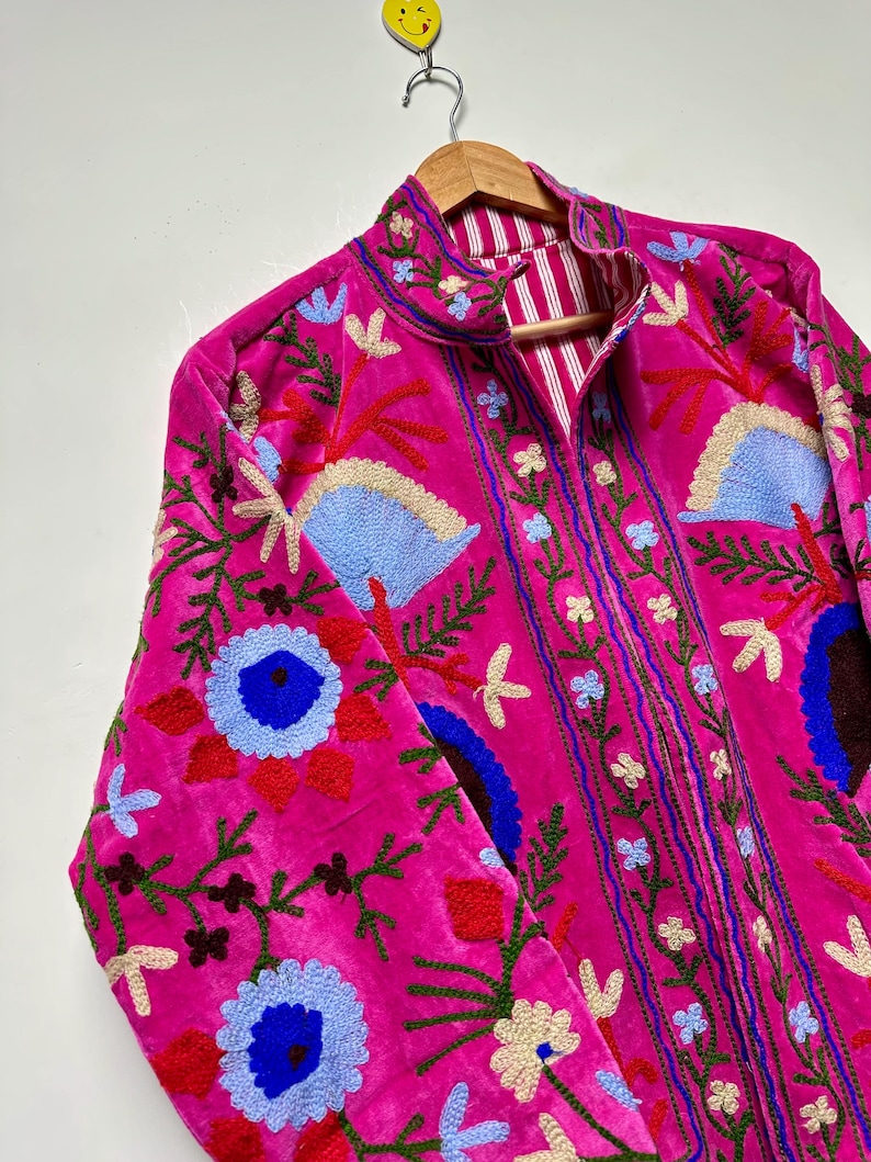 K&ouml;nnte beinhalten: Eine leuchtend pinke Samtjacke mit aufwendigen, mehrfarbigen Blumenstickereien in Blau-, Rot-, Gr&uuml;n- und Cremet&ouml;nen. Die Jacke hat einen Stehkragen, lange &Auml;rmel und ein sichtbares Innenfutter mit einem wei&szlig;-pinken vertikalen Streifenmuster.