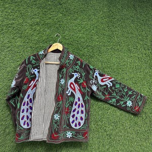 May include: A brown embroidered jacket with peacock and floral designs. The jacket features a striped lining and is displayed on a wooden hanger. The embroidery includes red, blue, and green accents, creating a vibrant and detailed pattern.