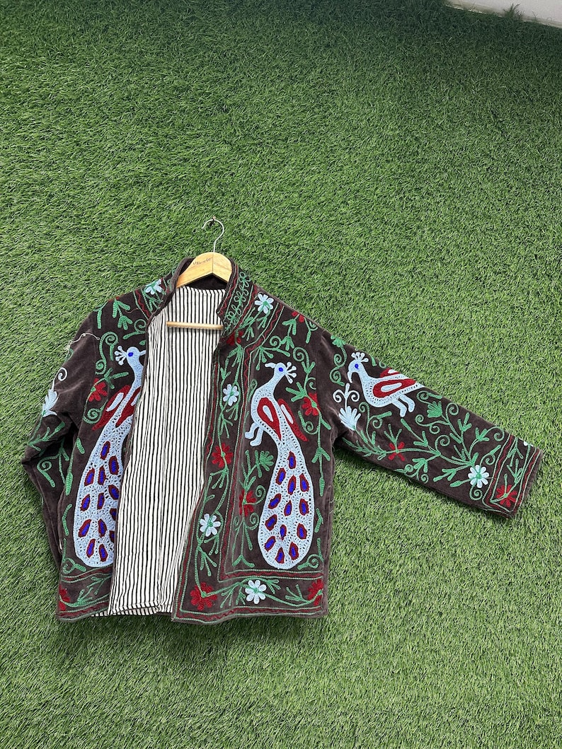 May include: A brown embroidered jacket with peacock and floral designs. The jacket features a striped lining and is displayed on a wooden hanger. The embroidery includes red, blue, and green accents, creating a vibrant and detailed pattern.
