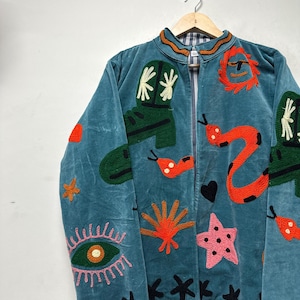 May include: Teal jacket with embroidered designs. Features green cowboy boots, orange snakes, a pink starfish, and black stars. The jacket has a zip-up front and a patterned collar. The jacket is hanging on a wooden hanger.