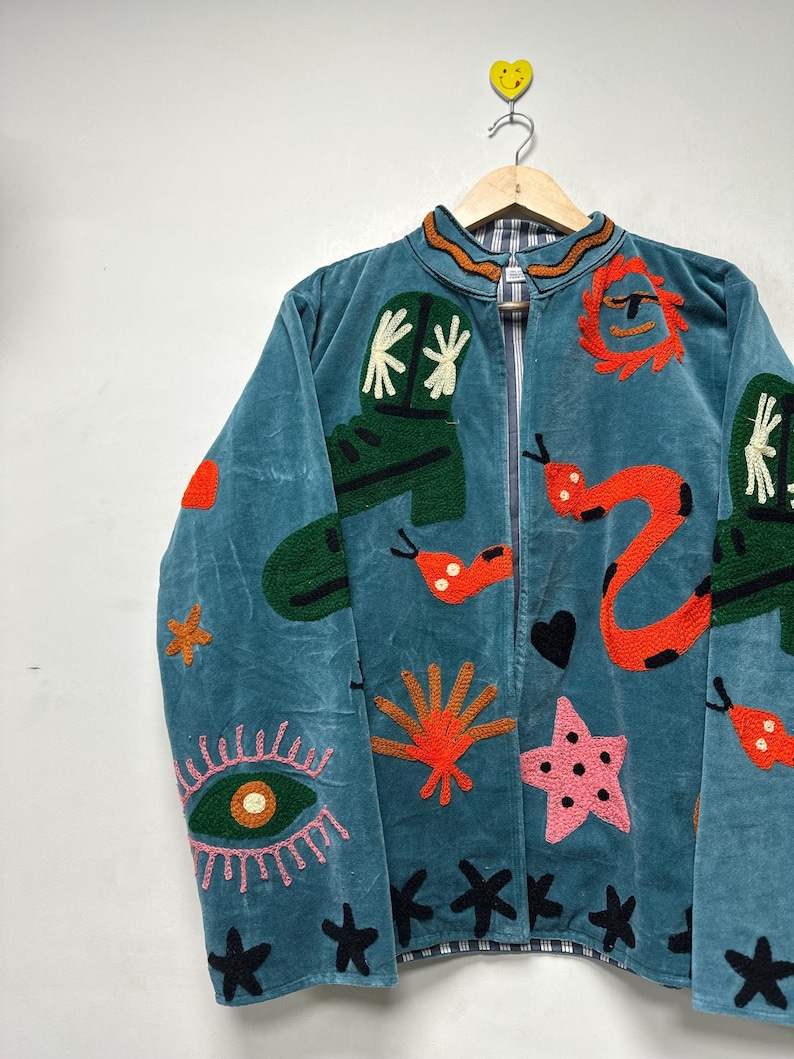 May include: Teal jacket with embroidered designs. Features green cowboy boots, orange snakes, a pink starfish, and black stars. The jacket has a zip-up front and a patterned collar. The jacket is hanging on a wooden hanger.