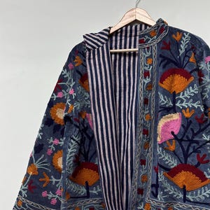 May include: A long-sleeved jacket in a muted blue fabric featuring dense, colorful floral embroidery in shades of orange, pink, and dark red. The interior lining is visible, showcasing a pattern of thin, vertical navy blue and white stripes.