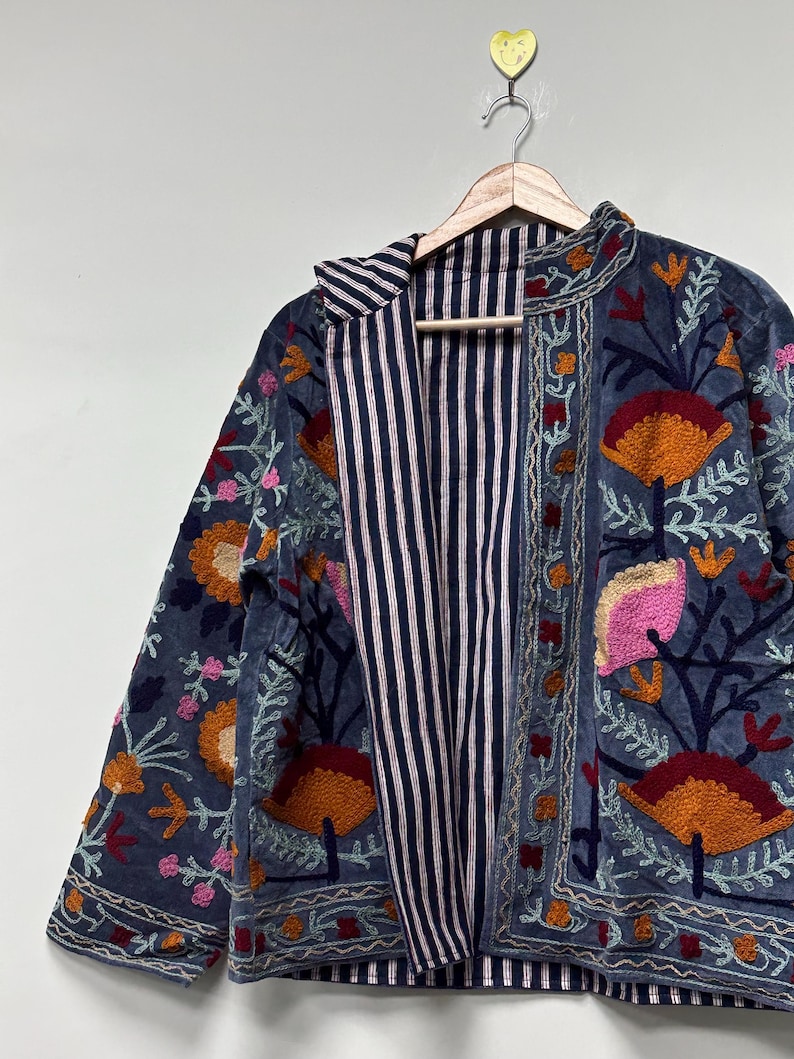 May include: A long-sleeved jacket in a muted blue fabric featuring dense, colorful floral embroidery in shades of orange, pink, and dark red. The interior lining is visible, showcasing a pattern of thin, vertical navy blue and white stripes.