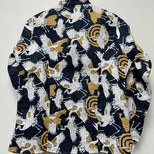 May include: A quilted jacket with a black background and a repeating pattern of white and gold cranes and circular designs. The jacket is hanging on a wooden hanger.