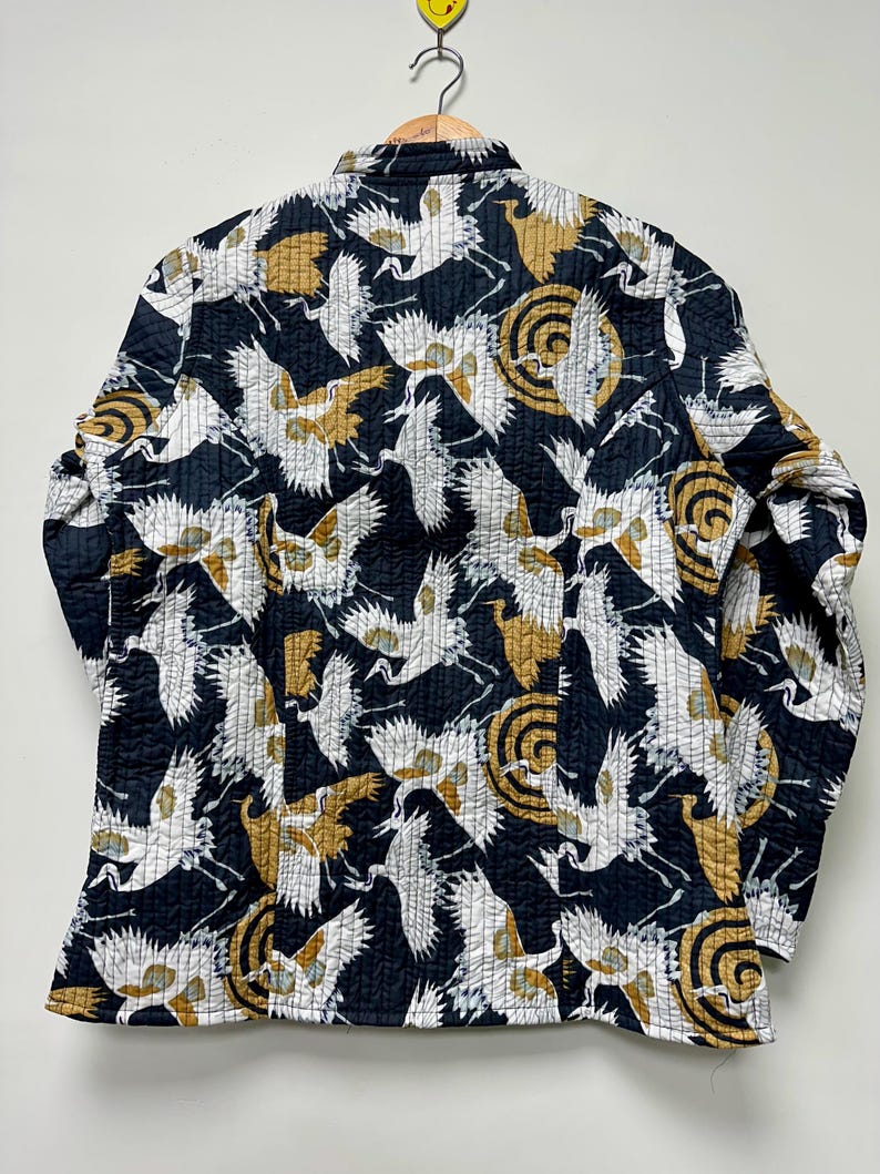 May include: A quilted jacket with a black background and a repeating pattern of white and gold cranes and circular designs. The jacket is hanging on a wooden hanger.