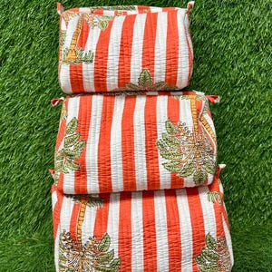 May include: Three quilted cosmetic bags stacked on top of each other. The bags have a pattern of vertical orange and white stripes. Each bag features a palm tree design with green and yellow leaves.