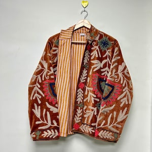 Puede incluir: Una chaqueta de terciopelo marr&oacute;n con intrincados bordados en crema, rojo y azul oscuro. La chaqueta tiene mangas largas, cuello y una parte delantera abierta que revela un forro de rayas verticales amarillas y blancas en el interior.