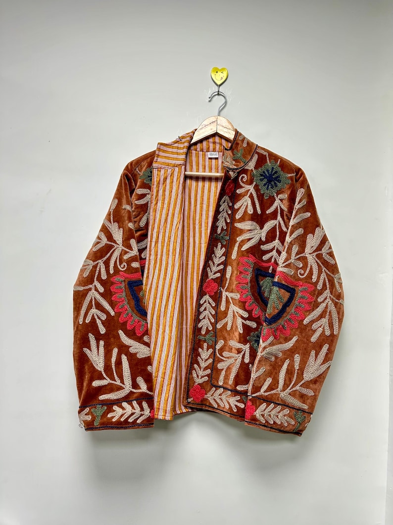 Puede incluir: Una chaqueta de terciopelo marr&oacute;n con intrincados bordados en crema, rojo y azul oscuro. La chaqueta tiene mangas largas, cuello y una parte delantera abierta que revela un forro de rayas verticales amarillas y blancas en el interior.