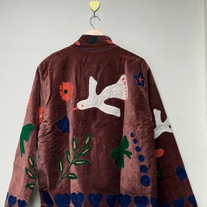 May include: A brown velvet jacket with embroidered details. The jacket features white birds, flowers, leaves, and blue hearts. The jacket is hanging on a wooden hanger against a white wall.