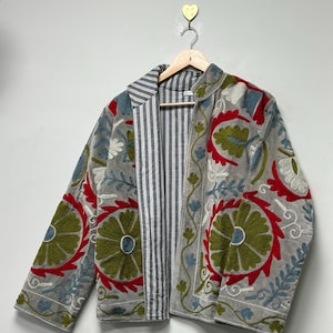 Può includere: Giacca grigia con ricami floreali intricati in rosso, verde e blu. La giacca presenta una fodera a righe ed è esposta su una gruccia di legno. Il ricamo crea un aspetto strutturato e decorativo.
