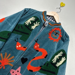 May include: Teal velvet jacket with colorful embroidered designs. Features include orange sun, red snakes, green shapes, and a pink star. The jacket is hanging on a wooden hanger with a yellow smiley face.