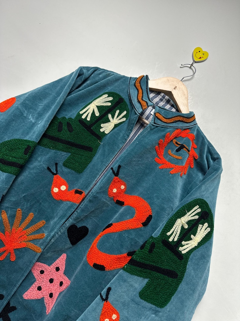May include: Teal velvet jacket with colorful embroidered designs. Features include orange sun, red snakes, green shapes, and a pink star. The jacket is hanging on a wooden hanger with a yellow smiley face.