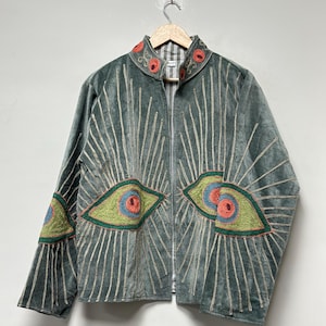 May include: A teal velvet jacket with a zip-up front, adorned with embroidered eye designs in green, orange, and blue. The jacket features vertical sequin lines radiating from the eyes and a collar with red floral embroidery.