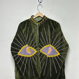 K&ouml;nnte beinhalten: Eine olivgr&uuml;ne Samtjacke mit Stehkragen und gelben, gestickten Blattmustern. Auf der Vorderseite befinden sich zwei gro&szlig;e, gestickte Augenmotive in Lavendel, Gelb und Rosa, die von strahlenf&ouml;rmigen, silbergrauen Sticklinien umgeben sind.