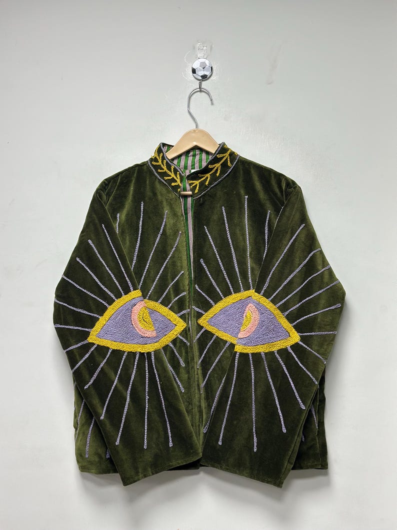 K&ouml;nnte beinhalten: Eine olivgr&uuml;ne Samtjacke mit Stehkragen und gelben, gestickten Blattmustern. Auf der Vorderseite befinden sich zwei gro&szlig;e, gestickte Augenmotive in Lavendel, Gelb und Rosa, die von strahlenf&ouml;rmigen, silbergrauen Sticklinien umgeben sind.