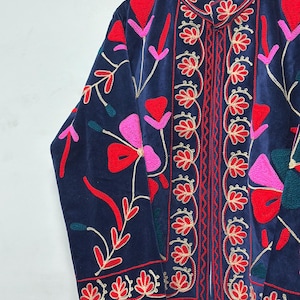 Op de afbeelding: Een marineblauw fluwelen jasje met levendig bloemenborduursel in rood-, roze- en blauwgroene tinten. Het kledingstuk heeft een opstaande kraag, lange mouwen en een opening aan de voorzijde met gedetailleerde rode stiksels en een gestreepte voering.