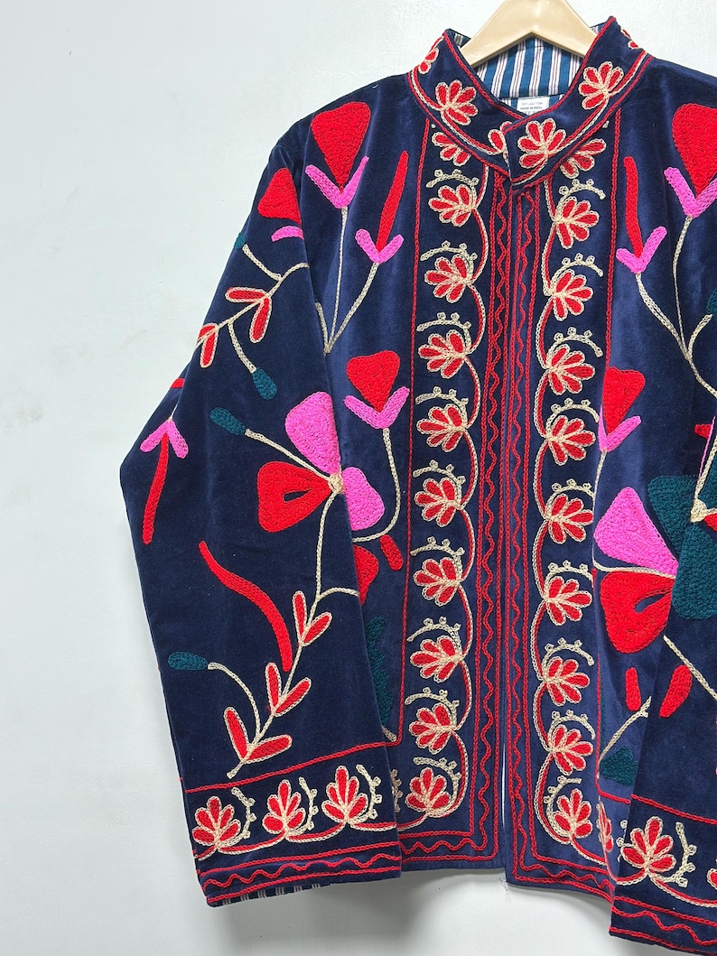 Op de afbeelding: Een marineblauw fluwelen jasje met levendig bloemenborduursel in rood-, roze- en blauwgroene tinten. Het kledingstuk heeft een opstaande kraag, lange mouwen en een opening aan de voorzijde met gedetailleerde rode stiksels en een gestreepte voering.