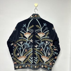 May include: A black velvet jacket with intricate floral embroidery in shades of beige, blue, and green. The jacket is hanging on a metal hanger against a white wall. The embroidery design is symmetrical, featuring stylized flowers and leaves.