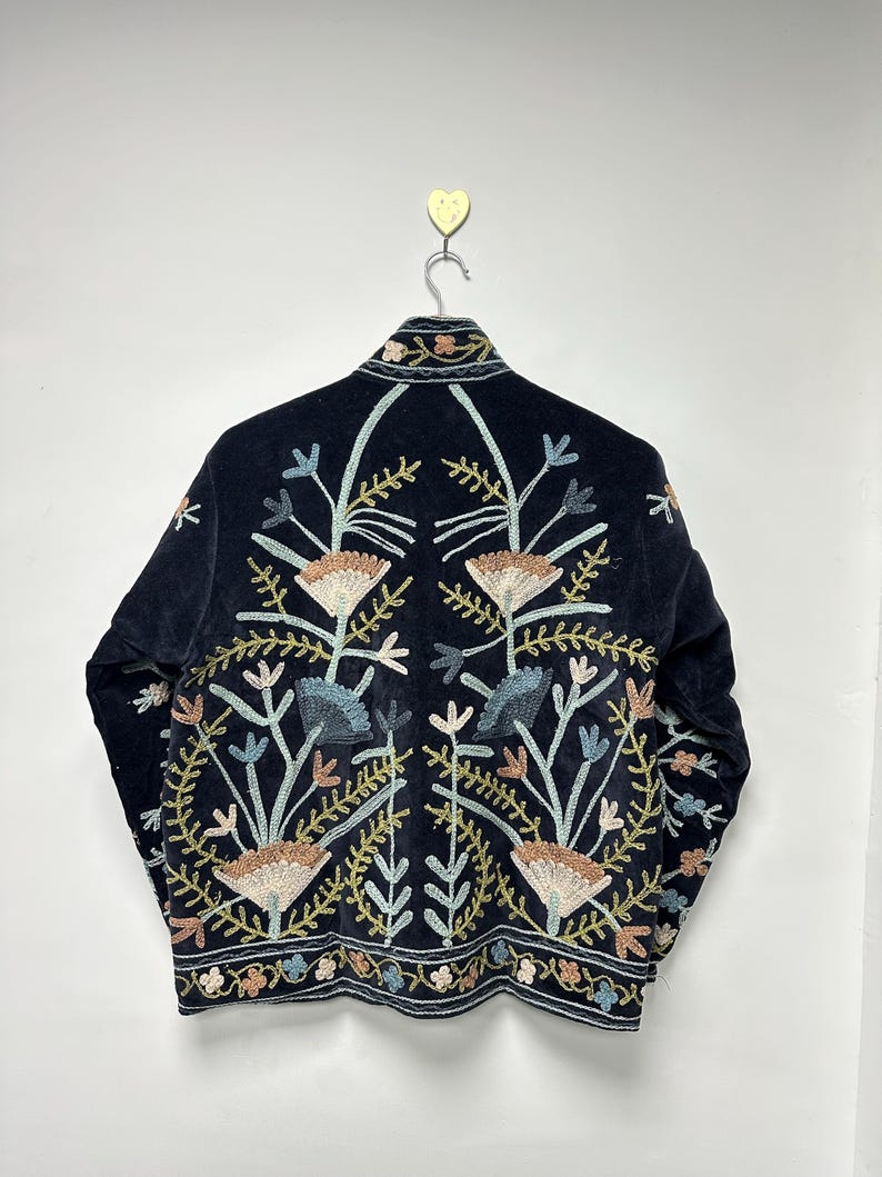 May include: A black velvet jacket with intricate floral embroidery in shades of beige, blue, and green. The jacket is hanging on a metal hanger against a white wall. The embroidery design is symmetrical, featuring stylized flowers and leaves.