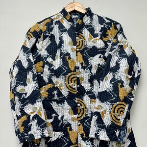 May include: A quilted jacket with a black background and a pattern of white and gold cranes. The jacket has a mandarin collar and is buttoned down the front. The jacket is hanging on a wooden hanger.