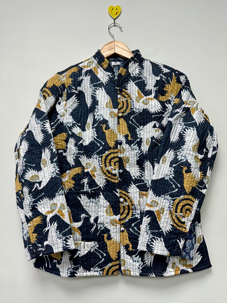 May include: A quilted jacket with a black background and a pattern of white and gold cranes. The jacket has a mandarin collar and is buttoned down the front. The jacket is hanging on a wooden hanger.
