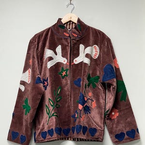 May include: A brown velvet jacket with a high collar and a zipper closure. The jacket is adorned with embroidered white birds, green leaves, blue hearts, and red and blue flowers. The jacket is hanging on a wooden hanger.