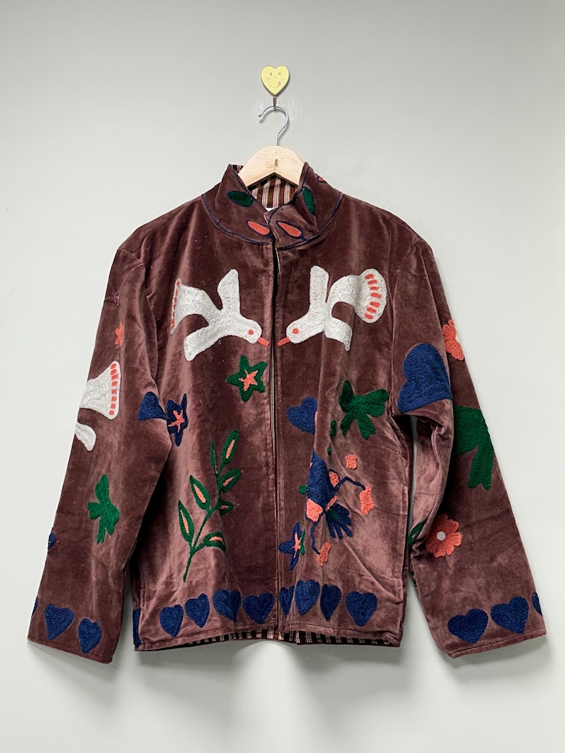 May include: A brown velvet jacket with a high collar and a zipper closure. The jacket is adorned with embroidered white birds, green leaves, blue hearts, and red and blue flowers. The jacket is hanging on a wooden hanger.