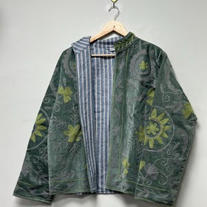 May include: A green velvet jacket with floral embroidery and a striped blue and white lining. The jacket is hanging on a wooden hanger against a white background. The embroidery features shades of green and gray.
