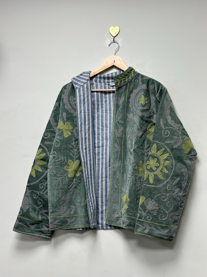 May include: A green velvet jacket with floral embroidery and a striped blue and white lining. The jacket is hanging on a wooden hanger against a white background. The embroidery features shades of green and gray.