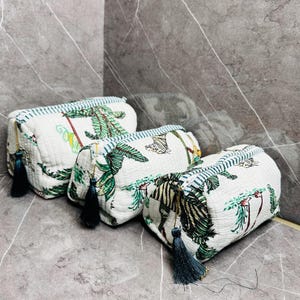 May include: Three white quilted makeup bags with a tropical print featuring palm trees, zebras, and butterflies. The bags have blue tassels and blue and white striped trim.