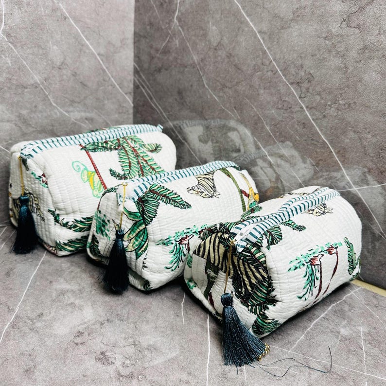 May include: Three white quilted makeup bags with a tropical print featuring palm trees, zebras, and butterflies. The bags have blue tassels and blue and white striped trim.