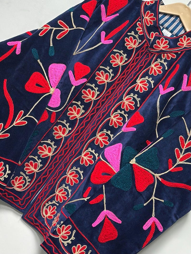 Op de afbeelding: Een marineblauw fluwelen jasje met levendig bloemenborduursel in tinten rood, roze en donkergroen. Het kledingstuk is voorzien van een mandarijnkraag, een knoopsluiting aan de voorzijde en een gestreepte voering zichtbaar bij de halslijn.