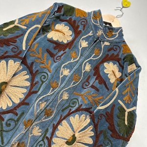 May include: A light blue velvet jacket featuring intricate floral embroidery in shades of cream, dark brown, and olive green. The garment has a high collar, a front zipper closure, and long sleeves, with dense botanical patterns covering the entire surface.