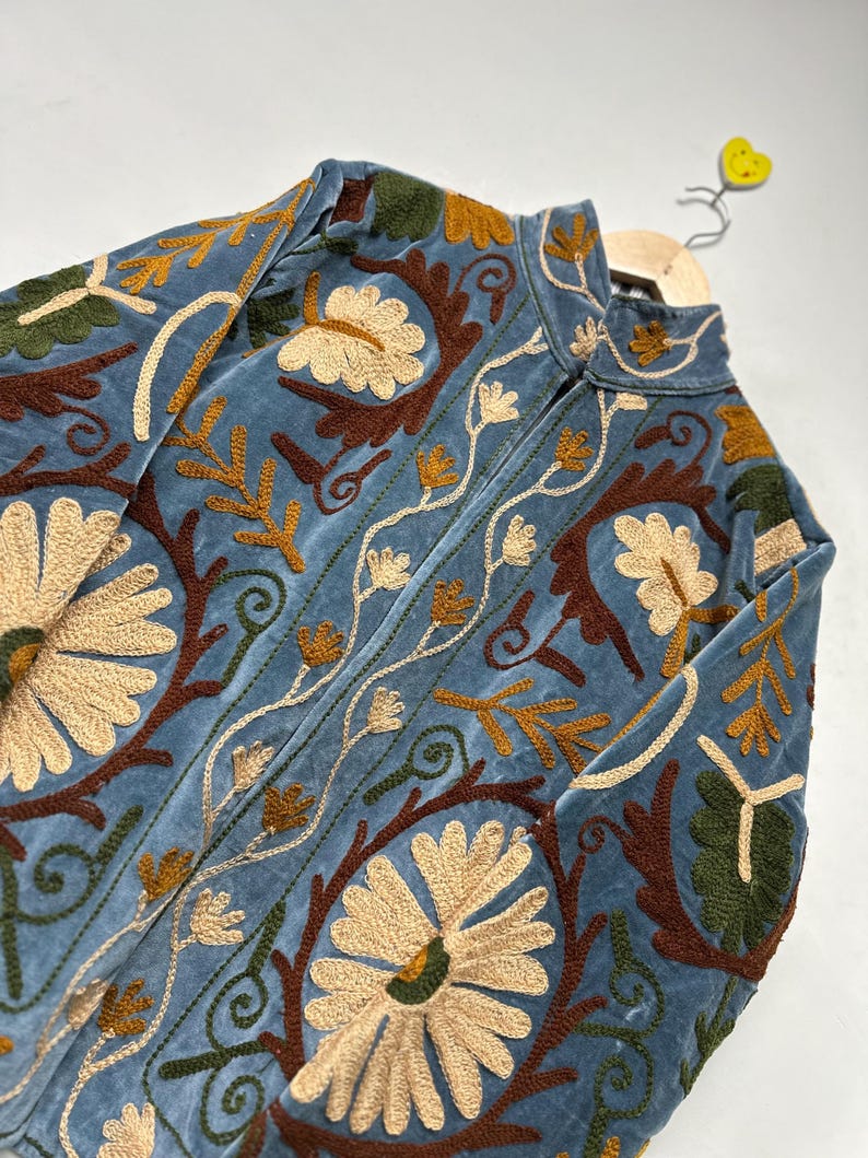 May include: A light blue velvet jacket featuring intricate floral embroidery in shades of cream, dark brown, and olive green. The garment has a high collar, a front zipper closure, and long sleeves, with dense botanical patterns covering the entire surface.